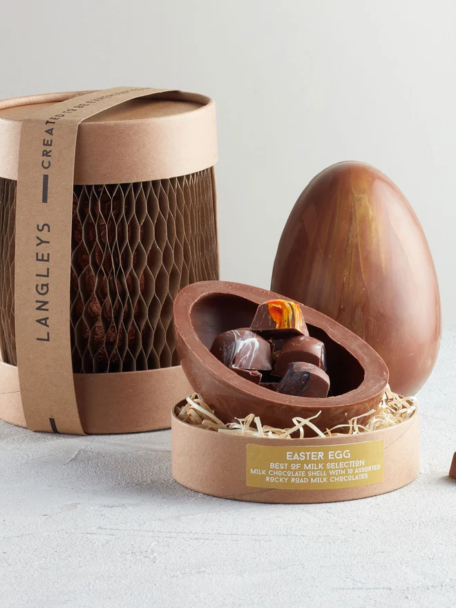 Best Of Milk Chocolate Easter Egg, a chocolate egg sitting on a white surface. It is accompanied by another chocolate egg that is opened showing the chocolates within. 