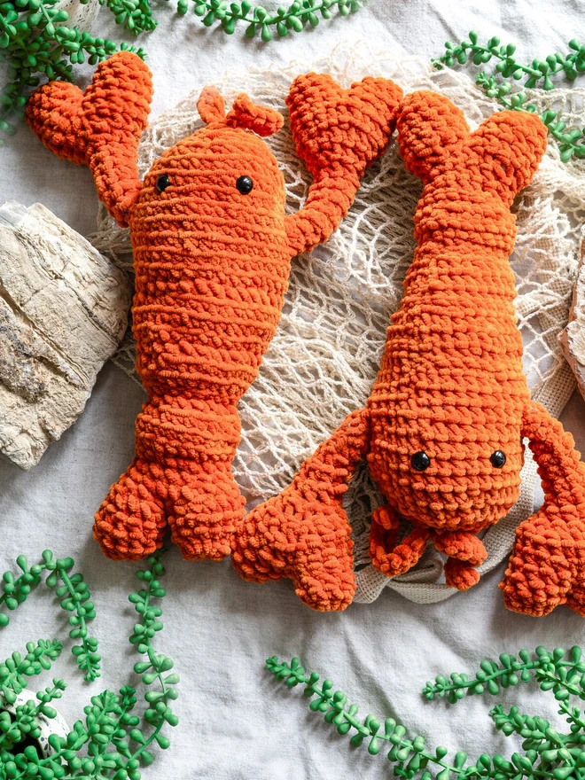 Chunky Chenille Cuddly Crochet Crustacean Lobster Plush, an orange lobster plush sitting next to another lobster on a white surface accompanied by a bag and some greenery. 