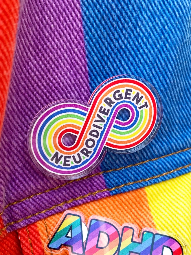 image shows an acrylic badge in the shape of a rainbow stripe infinity loop. printed across the loop is the word 'Neurodivergent'. the badge is pinned to a rainbow striped denim jacket.