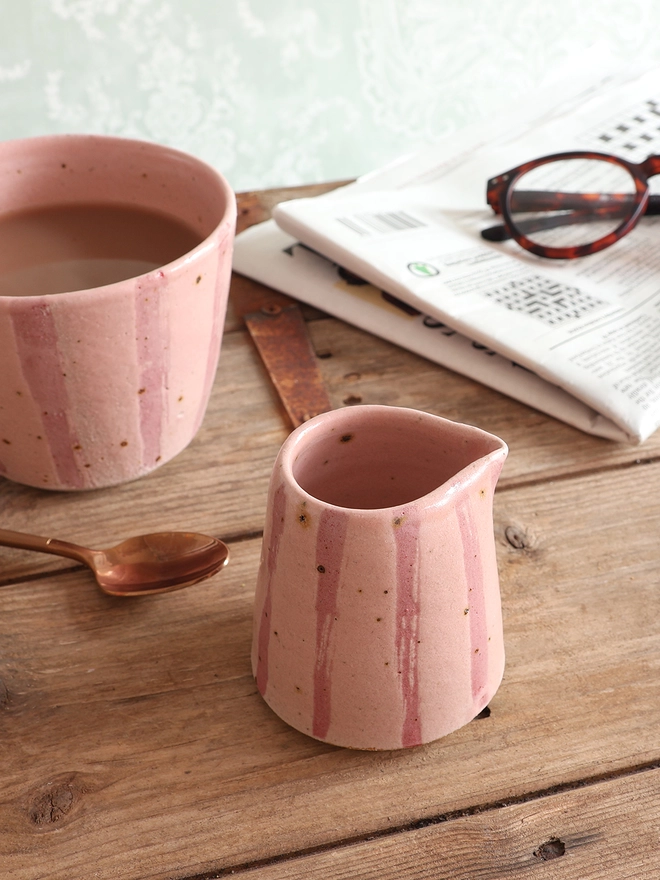 Handmade Ceramic Stoneware Small Jug in Pink Stripe sat on a wooden surface. A mug in the same style sits behind the mug, alongside a brass spoon and a newspaper with a pair of glasses on top. 