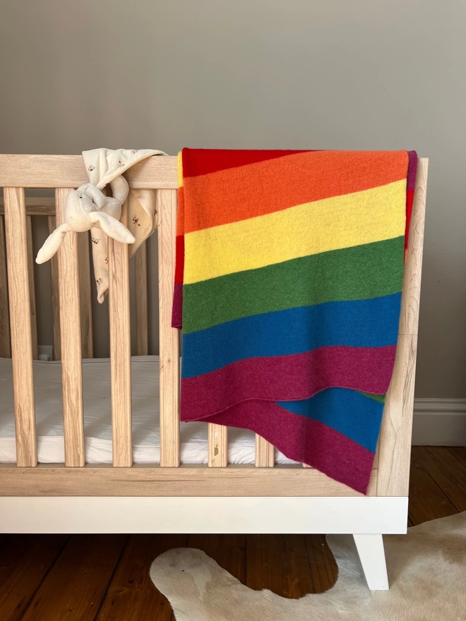 Knitted rainbow blanket shown hanging on side of a babies cot