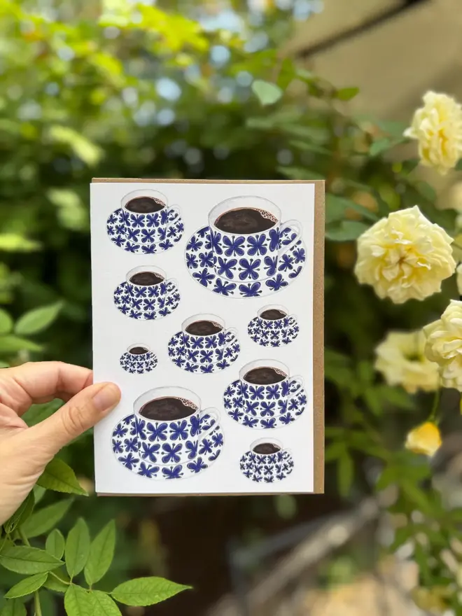 coffee cup and saucer illustrated card. card photographed against rose bush backdrop.