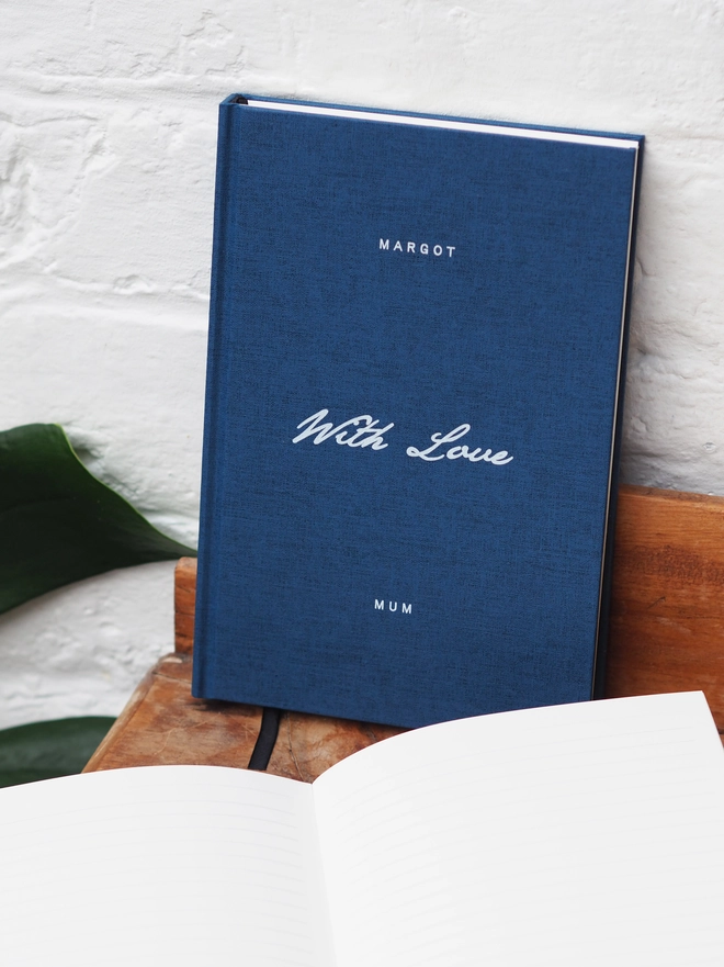 Personalised Hardcover With Love Notebook. A navy linen hardcover notebook with with words With Love written across the middle in contrasting white ink and personalised with a name at the top and bottom of the book. 