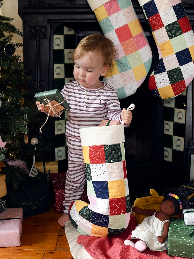 A toddler in striped pyjamas holds a patchwork quilted stocking in one hand and a small present in another.