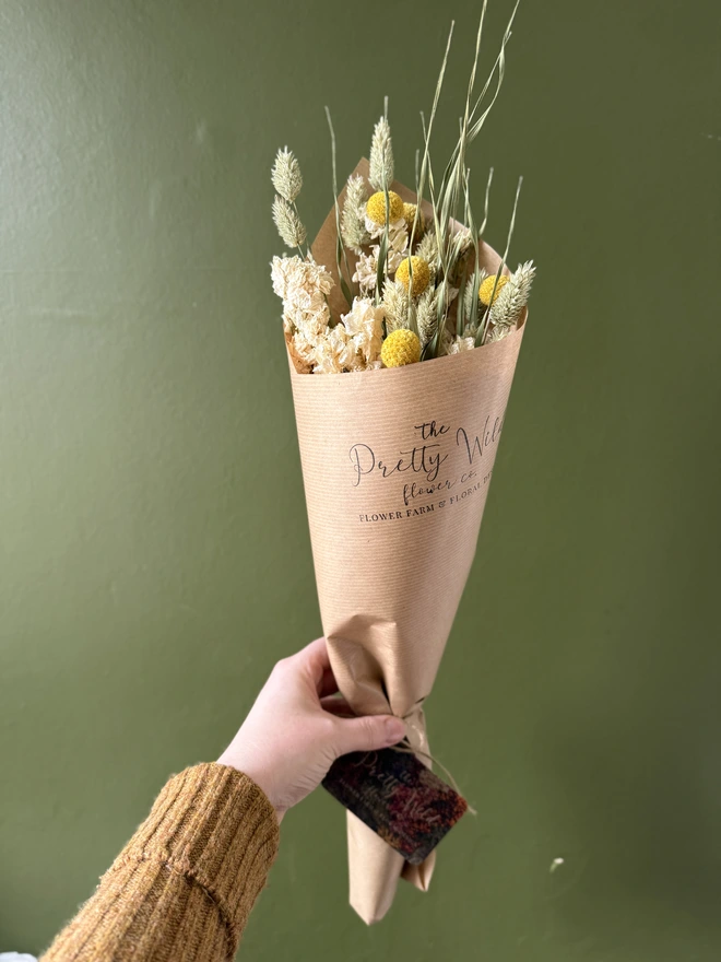 a bunch of dried flowers in bright colours wrapped in black tissue and kraft paper held by a person in a mustard coloured jumper with their left arm against a green wall