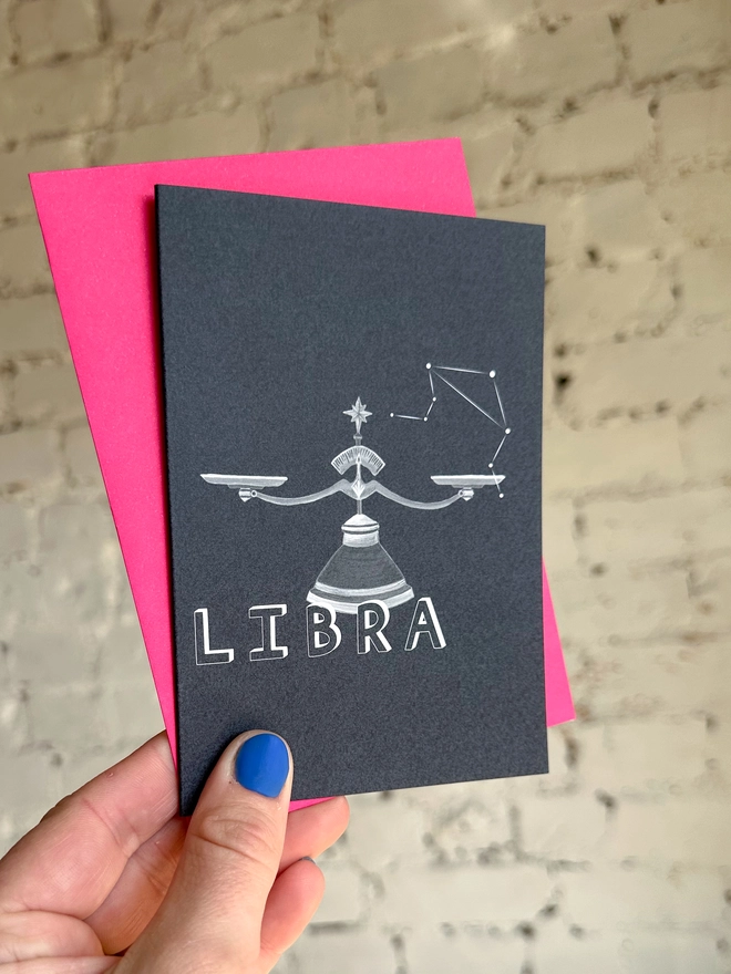  a greetings card featuring a greyscale illustration of a zodiac style set of scales with the text “LIBRA” with a neon pink envelope