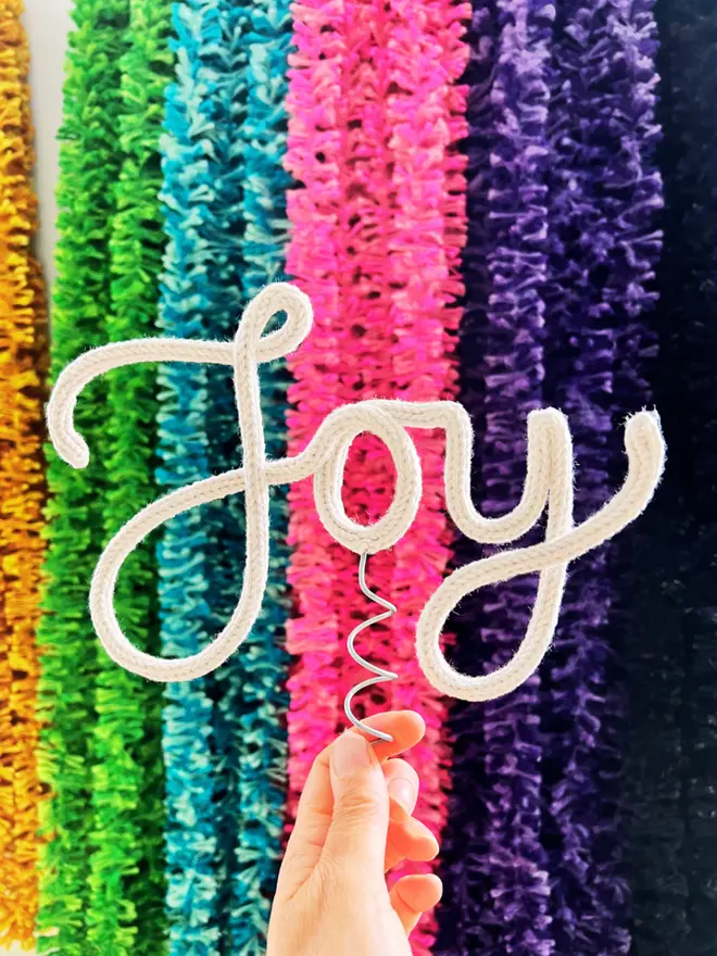 A handmade cotton string tree topper, spelling out the word 'Joy' in front of a display of multicoloured strinsel, held up by a hand