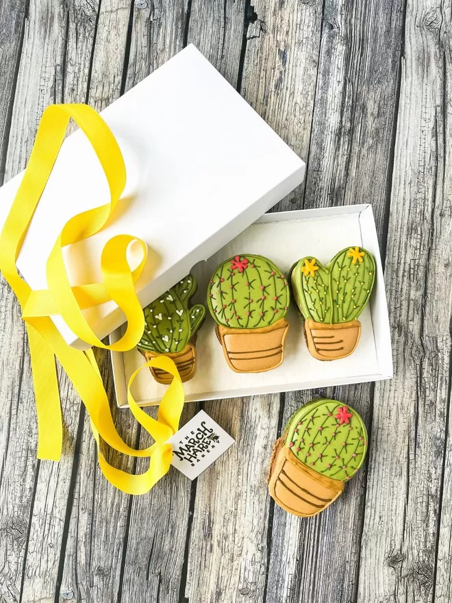 three large cactus shaped macarons decorated with white, yellow & pink flowers, in a white box with a yellow ribbon on a wooden table