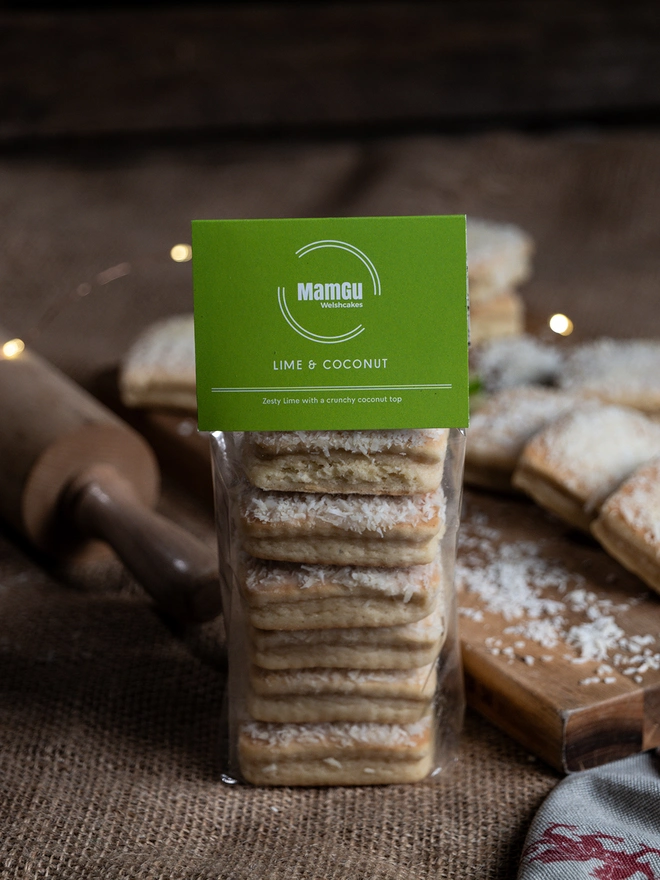 6 lime and coconut welsh cakes in a clear pack with a green mamgu welshcakes label on top, sitting on a messy rustic kitchen worktop