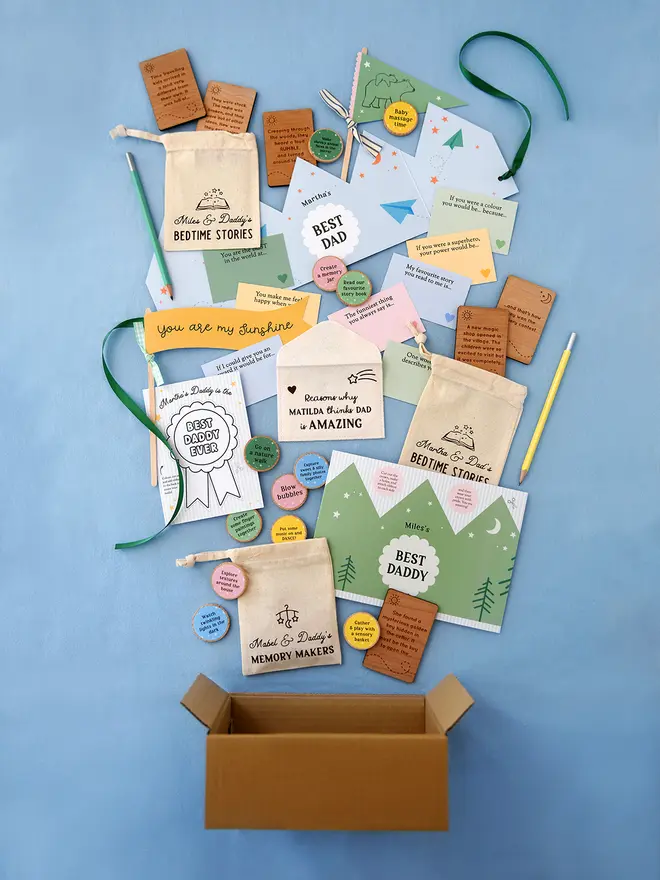 Flat lay of Clara & Macy’s heartfelt Father's Day gift collection for dads, featuring personalised bedtime story bags, wooden memory-making tokens, prompt cards, and keepsake envelopes from young children and babies, all styled on a soft blue background.