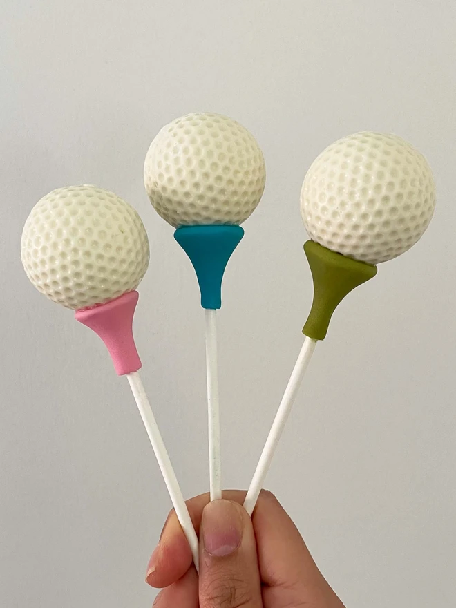 Golf Ball Cake Pops (Pack Of 6, 12, 18, 24), 3 cake pops placed in a hand against a white background. Each tee has it's own colour. 