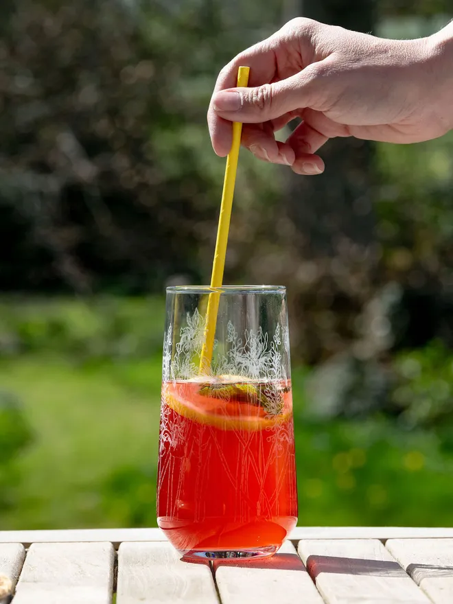 A hand placing a yellow straw into a tall, floral-etched highball glass filled with a red drink on an outdoor table.