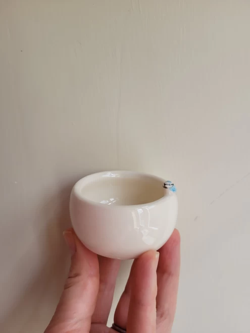 Hand holding a small off white ceramic tea light candle holder pot with a small modelled blue tit attached on the edge. the background is neutral