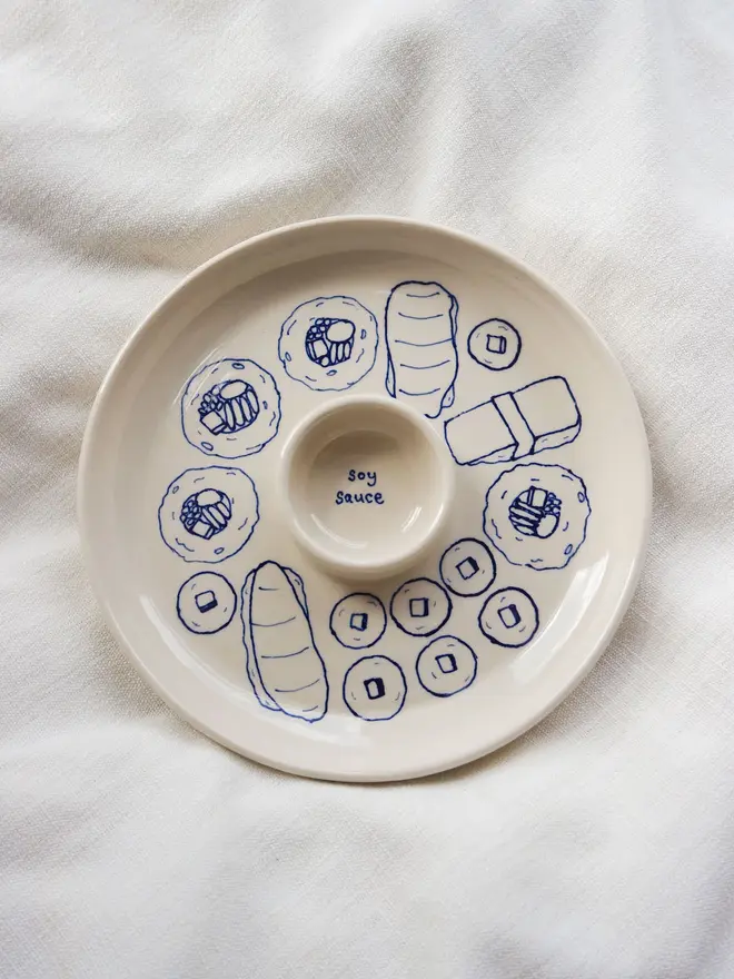 Snack Sushi Dippy Plate, a sushi snack dish sitting on a white surface. The outlines of sushi can be seen in blue. 