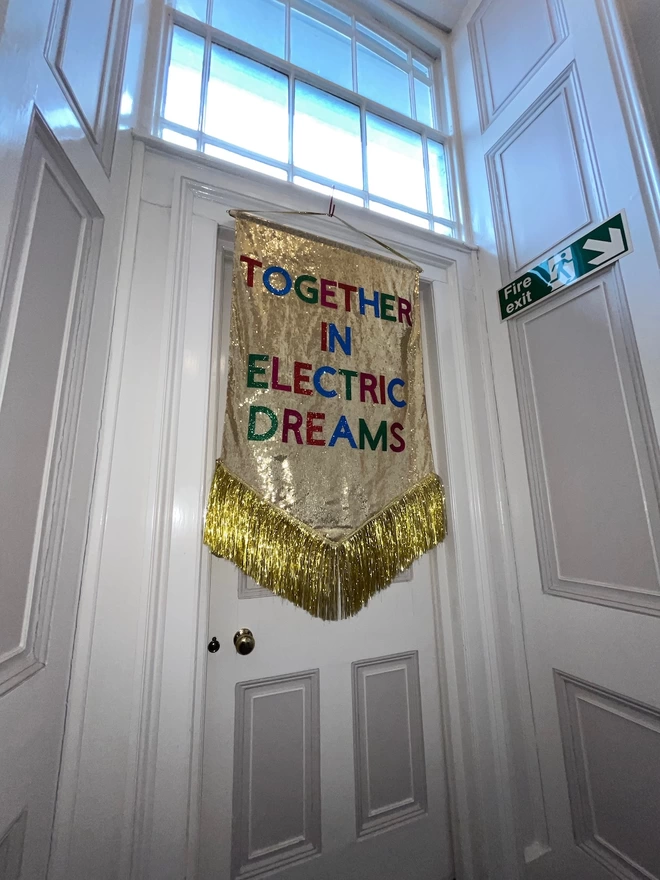 A maxi customisable banner hangs from a hook. It has a gold sequin background and a gold tinsel trim along the bottom. The text is multicoloured and says 'TOGETHER IN ELECTRIC DREAMS'