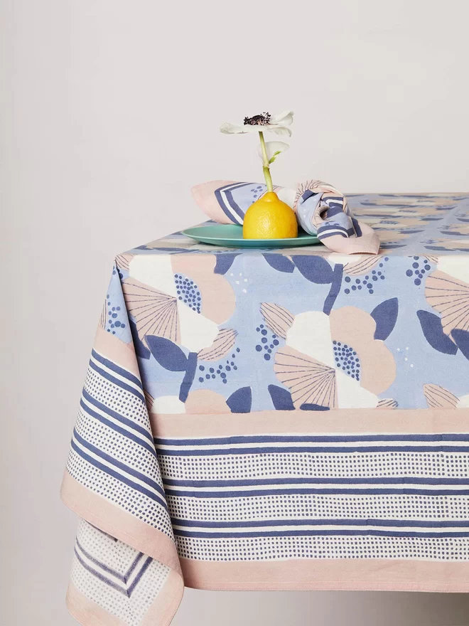 Blue block printed tablecloth featuring a large scale floral in cream, pink and navy colours with a soil pink border and navy stripe edging laid on a table