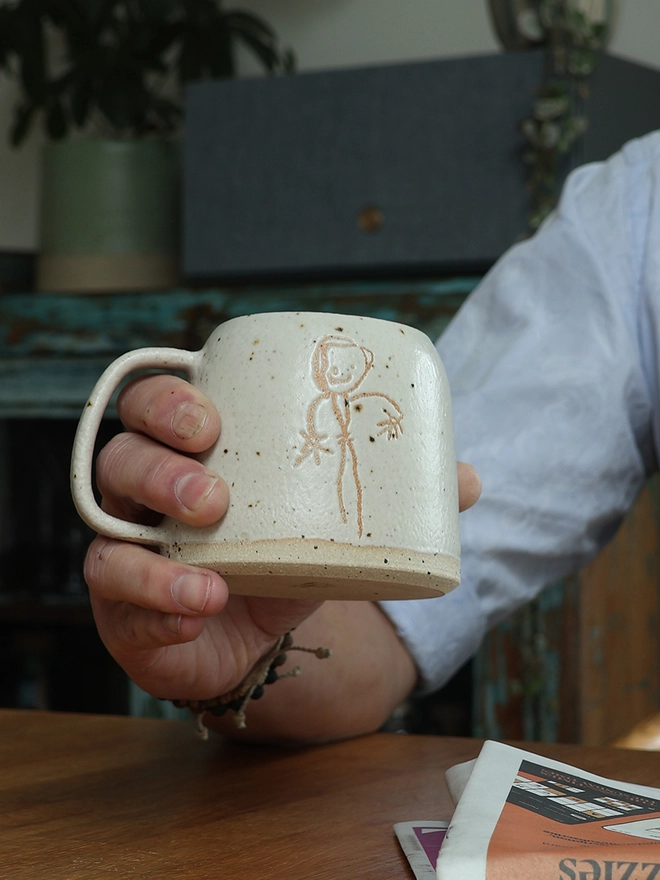 Childrens Drawing Handmade Ceramic Mug held in a hand over a wooden table. 