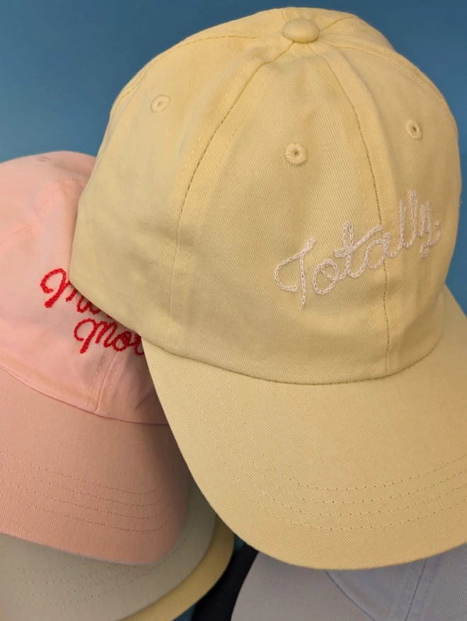 Yellow & Pink adult caps stacked on a blue background. The pink hat has red embroidery reading 'Moonlight Motel' & the yellow hat reads 'Totally' in white cursive embroidery