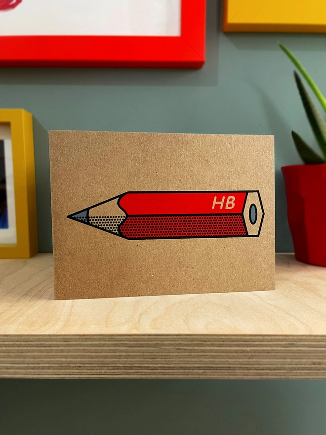 A red pencil design on brown Kraft card, with black outline and half tone detail. ON a plywood shelf with coloured picture frames around and a plant half in shot.