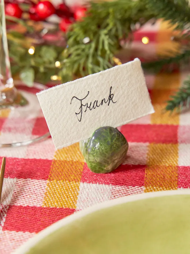A set of handmade ceramic Brussels sprout name card holders arranged on a yellow and red checkered tablecloth. Each glossy green sprout has individually modelled leafy details, and one is shown holding a heart-shaped display card.