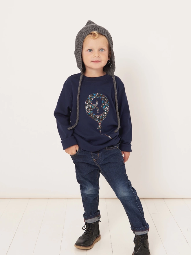 A smiling 3 year old boy wearing a navy cotton long sleeve t-shirt. The t-shirt features a balloon with the number 3 cut out from it, appliquéd in a starry Liberty print.