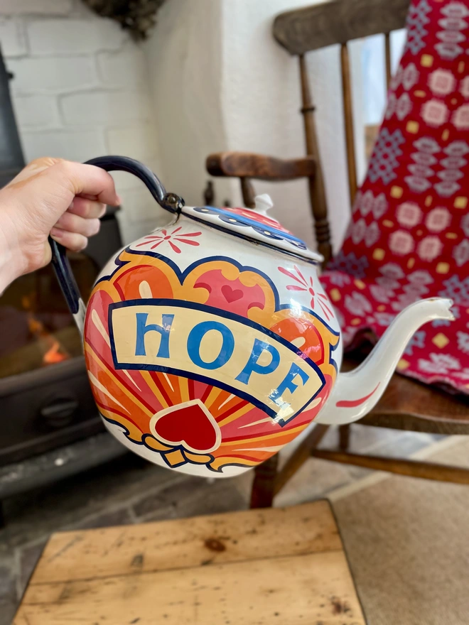 White vintage teapot, hand painted with a fairground inspired design in pink, orange, yellow and red. 'Hope' is painted in a banner across the centre.
