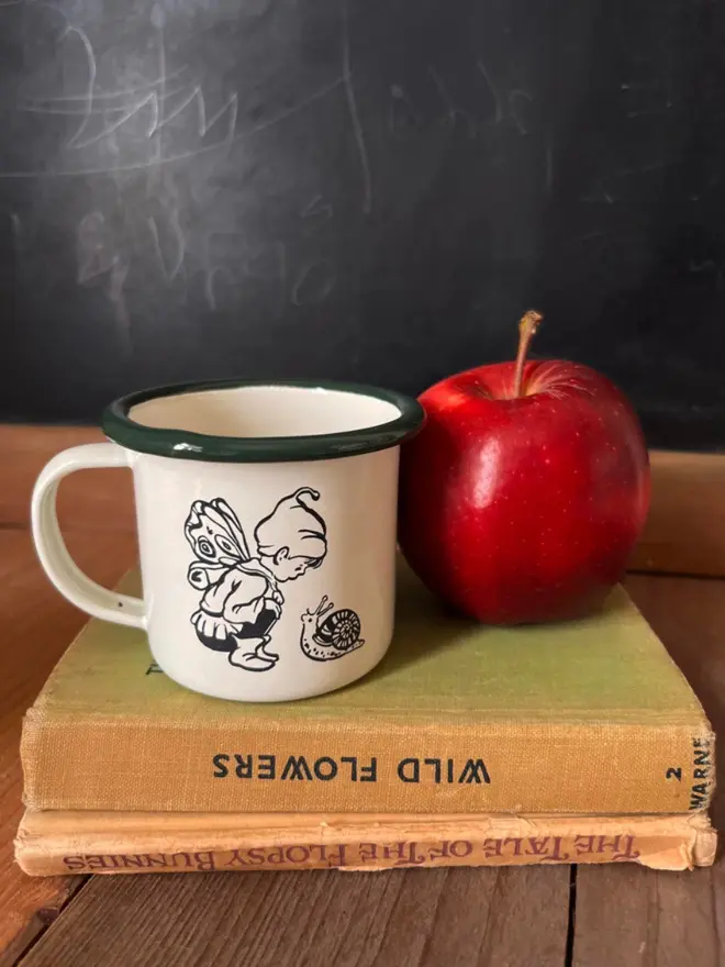a desk setting with stacked books, and a miniature enamel mug cream in colour with a bottle green rim featuring an illustration of a small snail and pixie