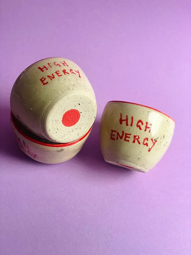 'High Energy' Mug,  a group of ceramic mugs that say 'High Energy' in front of a purple background. Each mug is stamped with a signature red dot on the bottom. 
