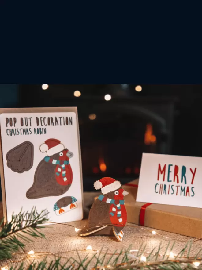 Robin Christmas decoration and Merry Christmas card on top of a hessian table cloth with fairy lights and a wood burning fire in the background