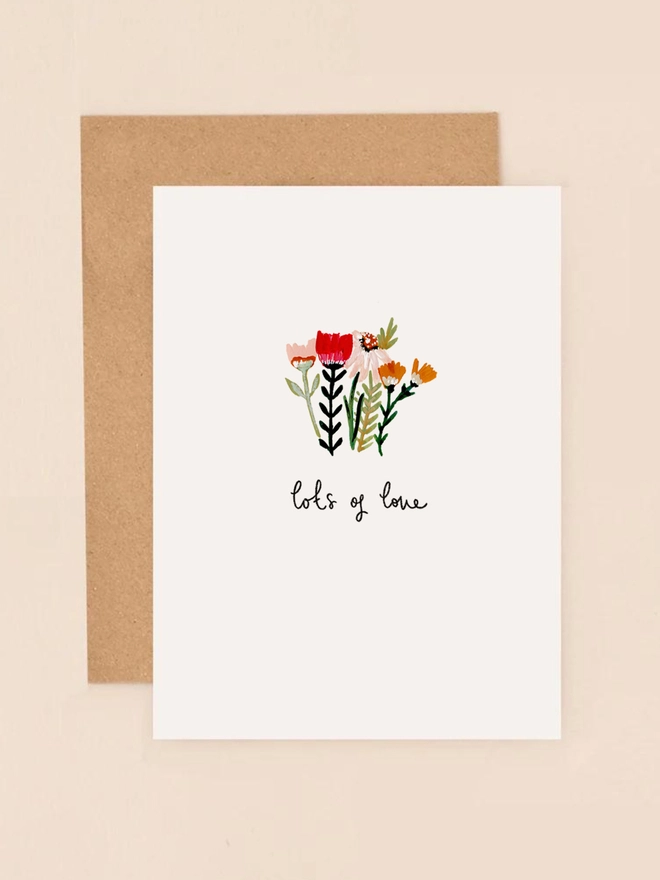A delicate illustration of a small bouquet featuring wildflowers in soft hues, including red, yellow, and green, arranged in a loose, hand-picked style. Beneath the flowers, the handwritten message "lots of love" adds a personal and heartfelt touch. The design is minimalist and charming, printed on thick, textured FSC-certified board
