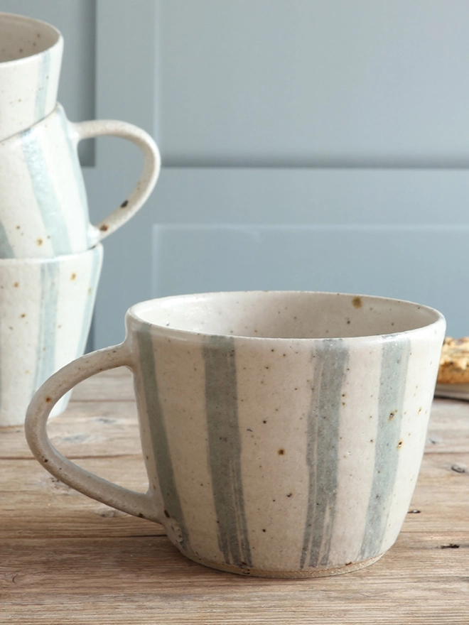 Handmade Ceramic Stoneware Mug in Blue and White Stripe sat on a wooden surface. The background shows three of these mugs stacked on top of each other. 