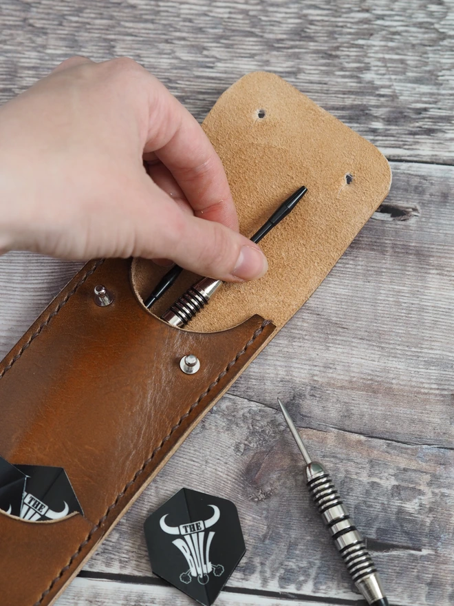 Handmade leather darts sleeve. A hand pulls out a single dart with flights scattered around ready to be assembled.