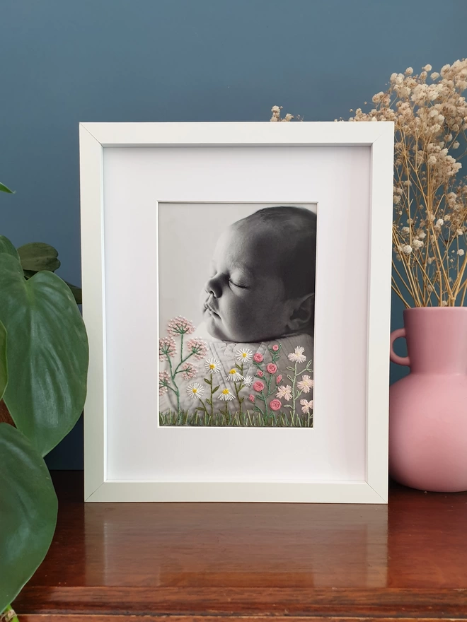 Baby photo with hand embroidered flowers growing from bottom of image, white frame on desk