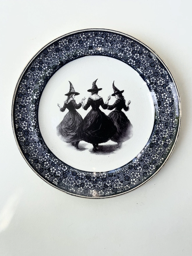 a coven of 3 witches dance hand in hand, the image is victorian in black and white and is printed directly onto an ornate vintage plate