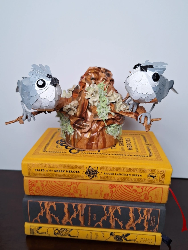 A handmade paper sculpture depicting two tufted titmice birds perched on a tree stump. The sculpture is placed on top of a pile of books.