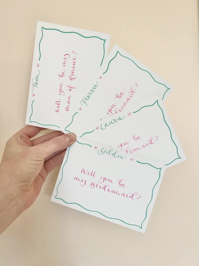 A6 notecards with a mint green wiggly border and a name inside the top border line, bookended by a pink heart either side. Pink handwritten text inside the border reads 'Will you be my bridesmaid?' and 'Will you be my Maid of Honour?'