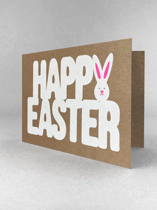 A slightly angled landscape kraft card on a grey background. The words Happy Easter are screen printed in white ink. The letter Y of happy forms the shape of a white bunny. Neon pink ink is printed over the top to create the ears and the classic twitchy bunny nose.