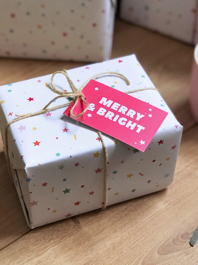 Gifts wrapped in pastel star Christmas wrapping paper are on a wooden floor beneath a Christmas Tree.