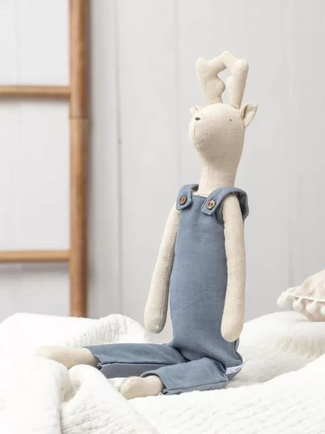 The Adventure Pickles Deer Soft Toy In Blue Dungarees Sitting On Bed