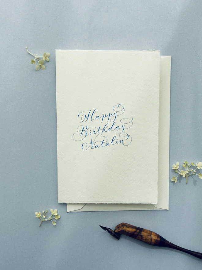 Blue calligraphy lettering on a handwritten birthday card next to a calligrapher's pen