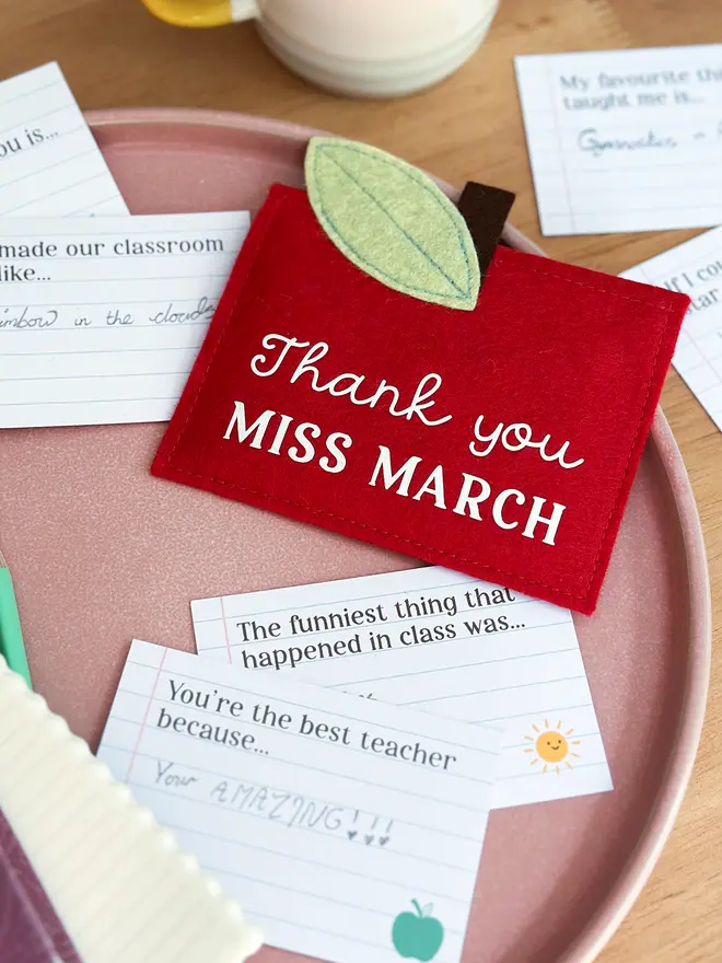 A handmade red felt apple-shaped envelope with "Thank you Miss March" written on the front. The heirloom apple envelope surrounded by completed prompt cards with handwritten messages from a child, displayed on a pink plate and wooden desk. It's a thoughtful personalised gift for teachers from children.