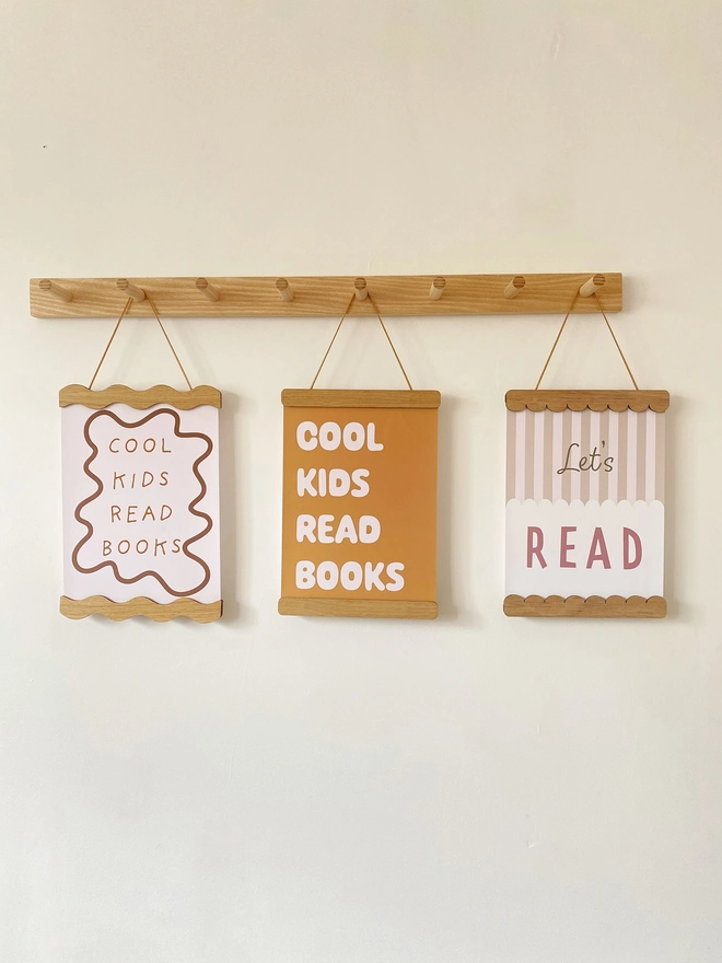 Three wooden picture frames hung on a peg rail. the picture frames are straight, wavy and scalloped in style. Each is strung with vegan suede and holds children's colourful prints reading Cool Kids Read Books and Lets Read