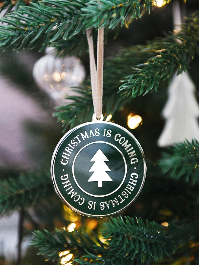 A deep green and gold enamel Christmas decoration, with the words Christmas Is Coming surrounding a gold Christmas tree, hangs on a Christmas tree.