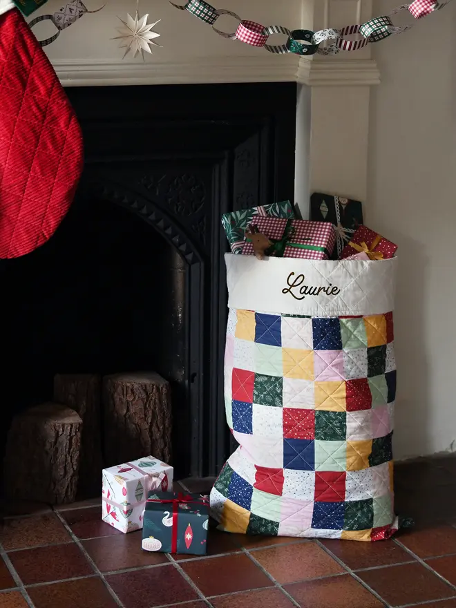 A handmade patchwork Christmas sack sits beside a black fireplace.