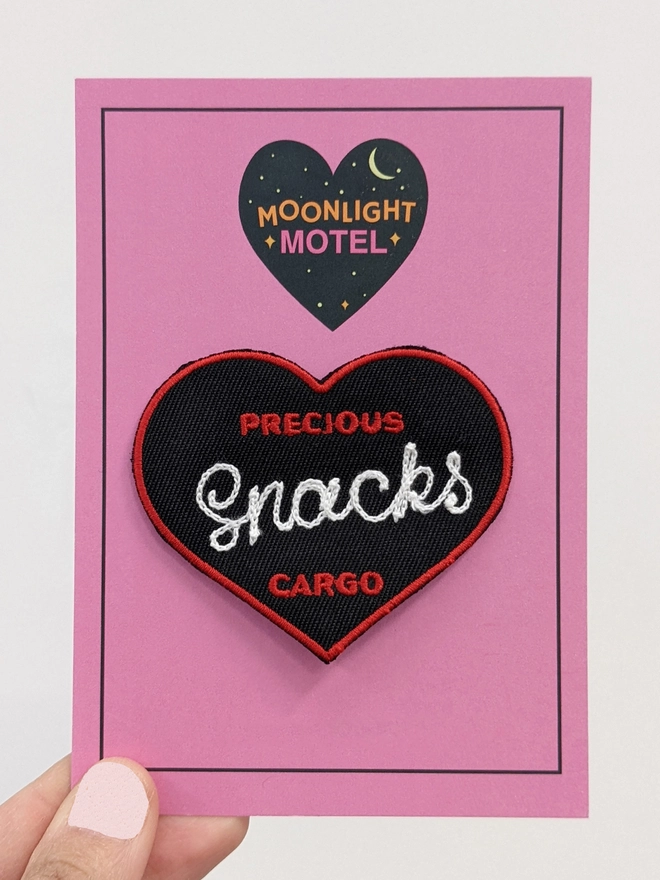 Black, Red & White Woven patch, customised with White Chainstitch embroidery reading 'Snacks'. The words 'Precious' above, & 'Cargo' below in Red. Presented on a pink Moonlight Motel backing card, and held up to a white background.  