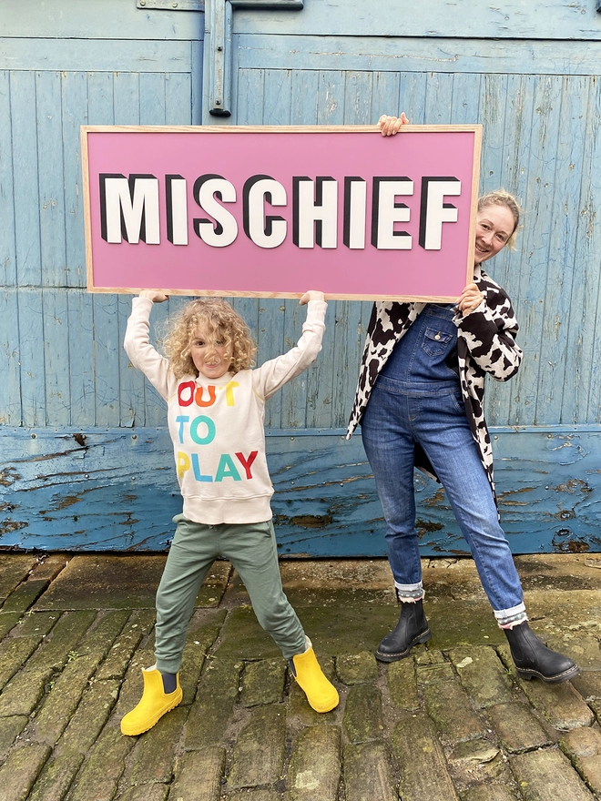 2 people holding a sign with a pink background up in the air which reads MISCHIEF 