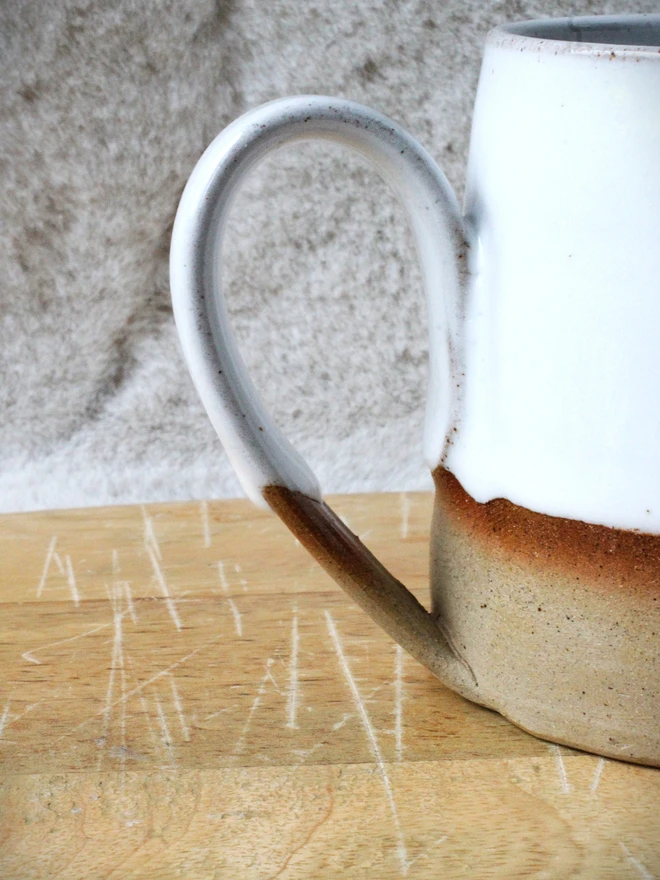 Rustic ceramic mug with a white speckled upper glaze and brown lower half, featuring the word “Brave” on the front, sitting on a wooden table against a neutral background.