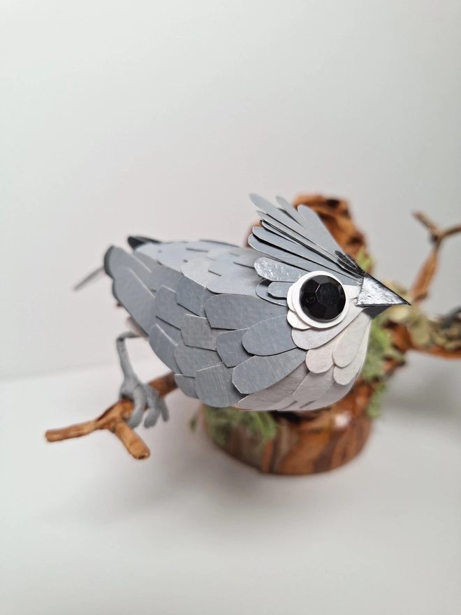 Tufted Titmice on a trees tump handmade bird sculpture. lose up of one titmouse perched on a brancg. The bird is made from grey and white paper feathers