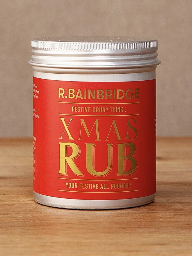 XMAS Rub Seasoning, a jar of Christmas seasoning sitting on a wooden surface in front of a plain backdrop. 