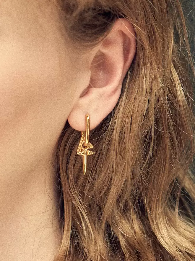 Woman wearing an elongated hoop styled with a perpetuity triangle charm and a small bayonet gold charm